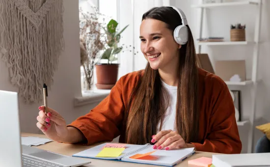 Estudiante sonriendo frente a su computador portátil con audífonos y apunte